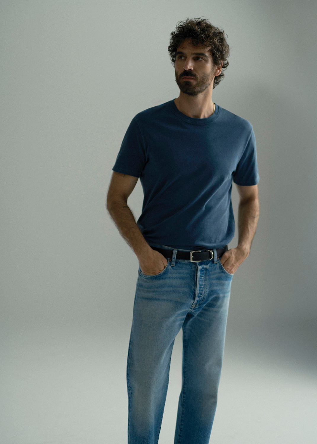 A man wearing a dark navy blue crew-neck t-shirt and light blue jeans, standing against a neutral gray studio background.