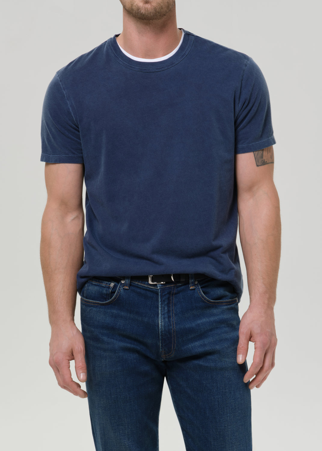 A man wearing a faded navy blue crewneck t-shirt, tucked into dark blue jeans, against a grey background.