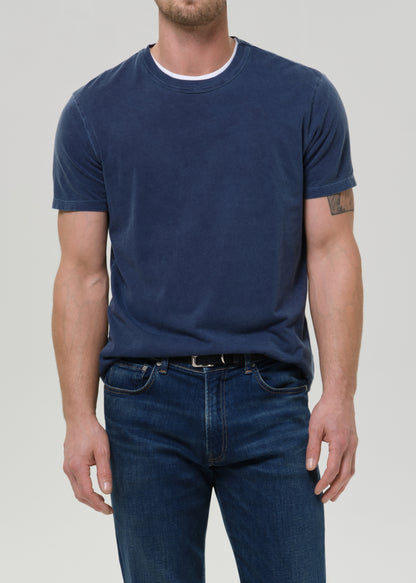A man wearing a faded navy blue crewneck t-shirt, tucked into dark blue jeans, against a grey background.