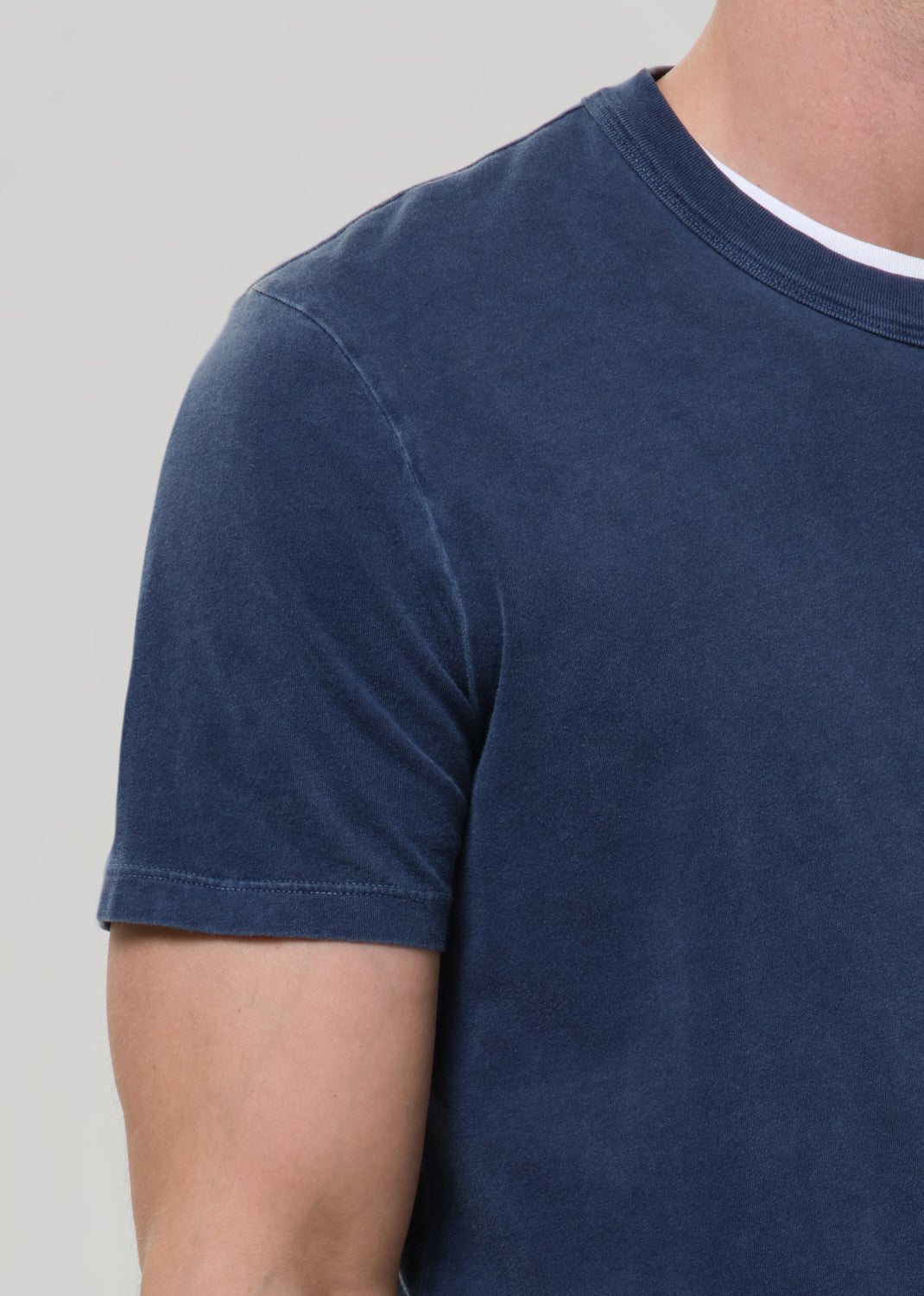 Close-up of a person wearing a short-sleeve crew-neck t-shirt in a washed navy blue color, layered over a white undershirt, against a light grey background.