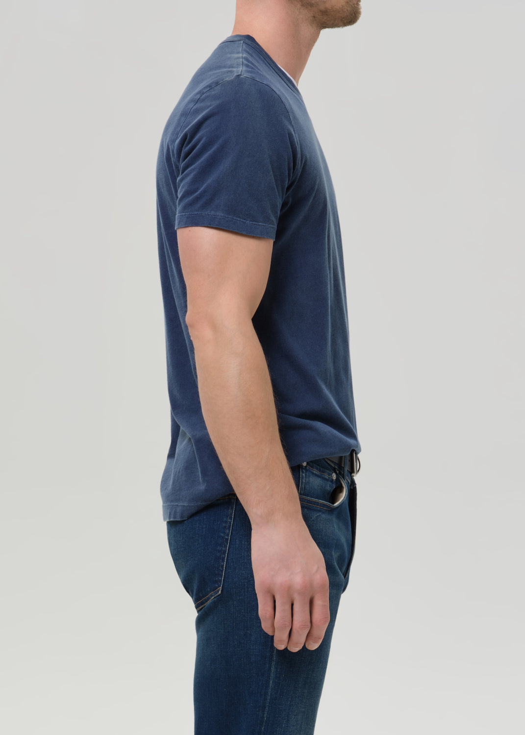 A man side shot wearing a faded navy blue crewneck t-shirt, tucked into dark blue jeans, against a grey background.