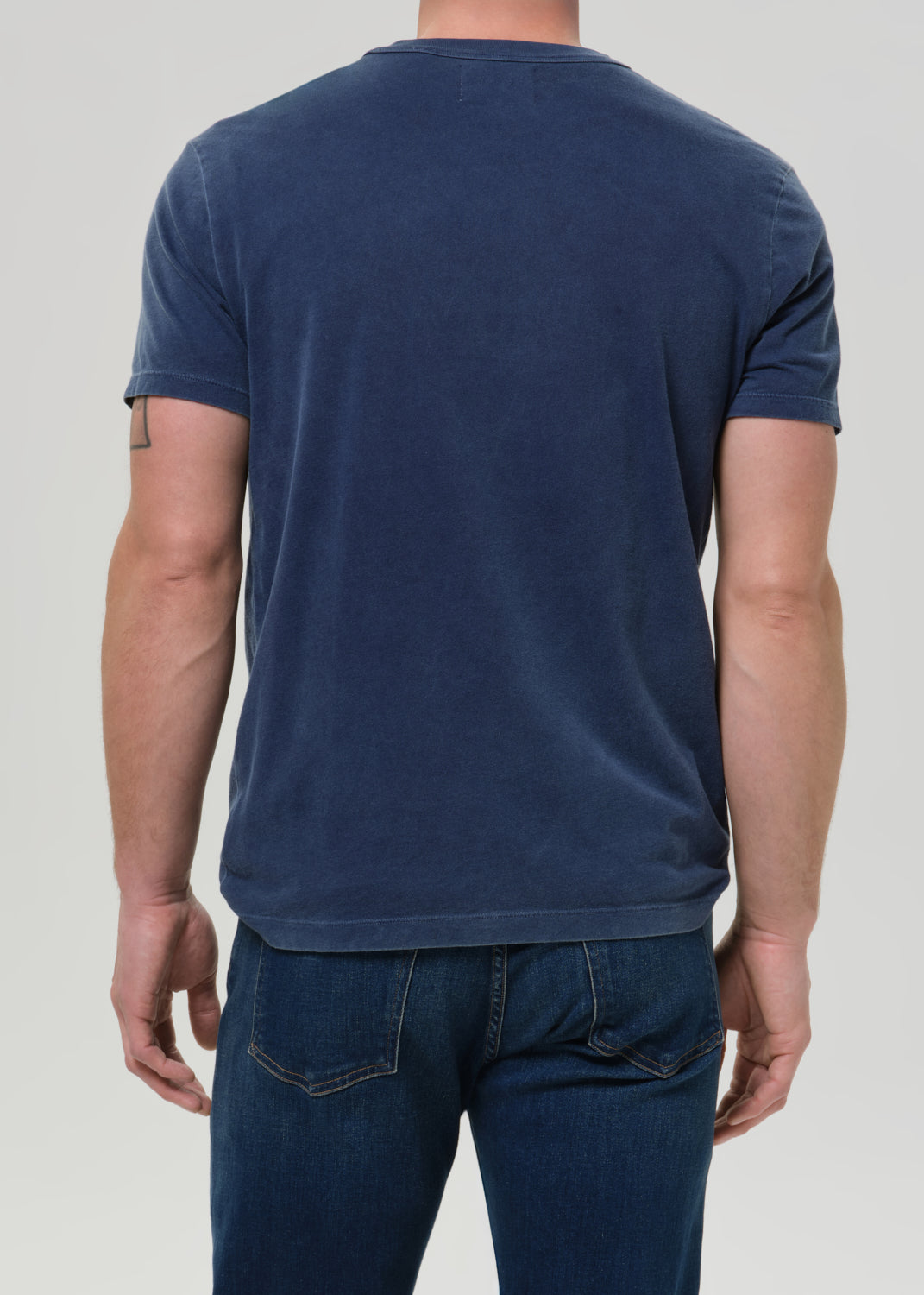 A man back shot wearing a faded navy blue crewneck t-shirt, tucked into dark blue jeans, against a grey background.