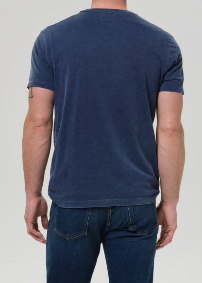 A man back shot wearing a faded navy blue crewneck t-shirt, tucked into dark blue jeans, against a grey background.