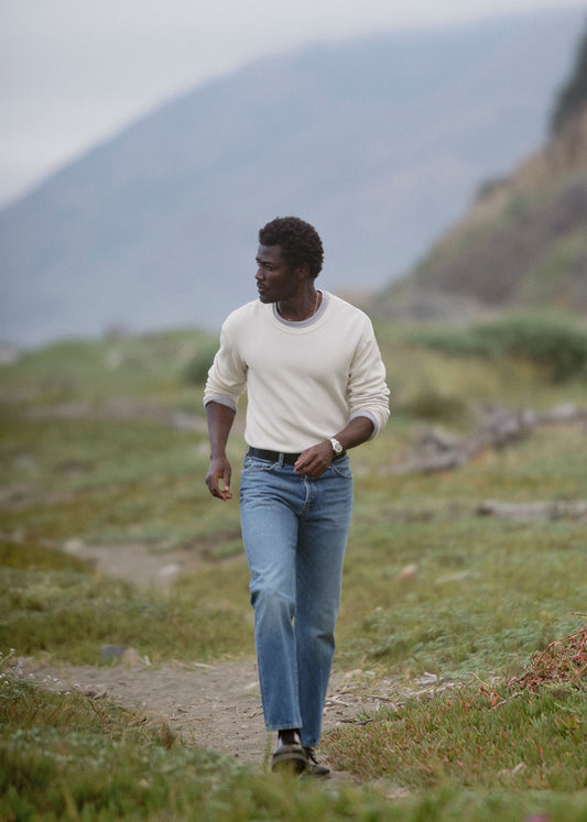 A man walking on a grassy path in an outdoor mountain setting, wearing a cream long-sleeve crewneck shirt and blue jeans.