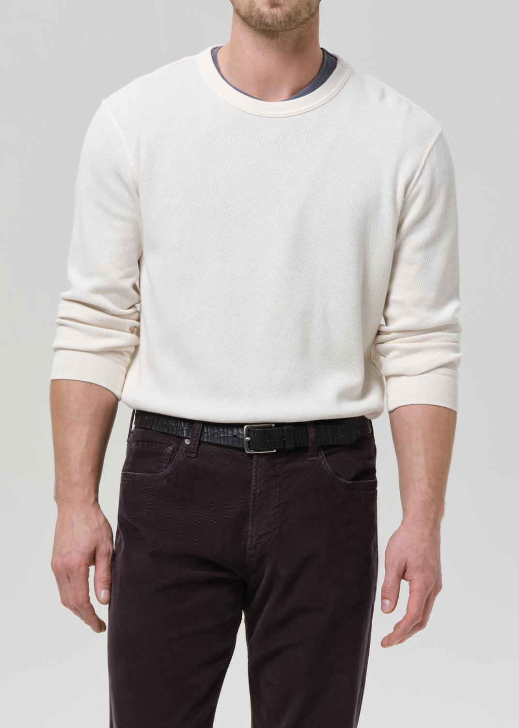 A man wearing a cream-colored long-sleeve crewneck sweater and dark charcoal pants, shown from the neck down against a grey background.