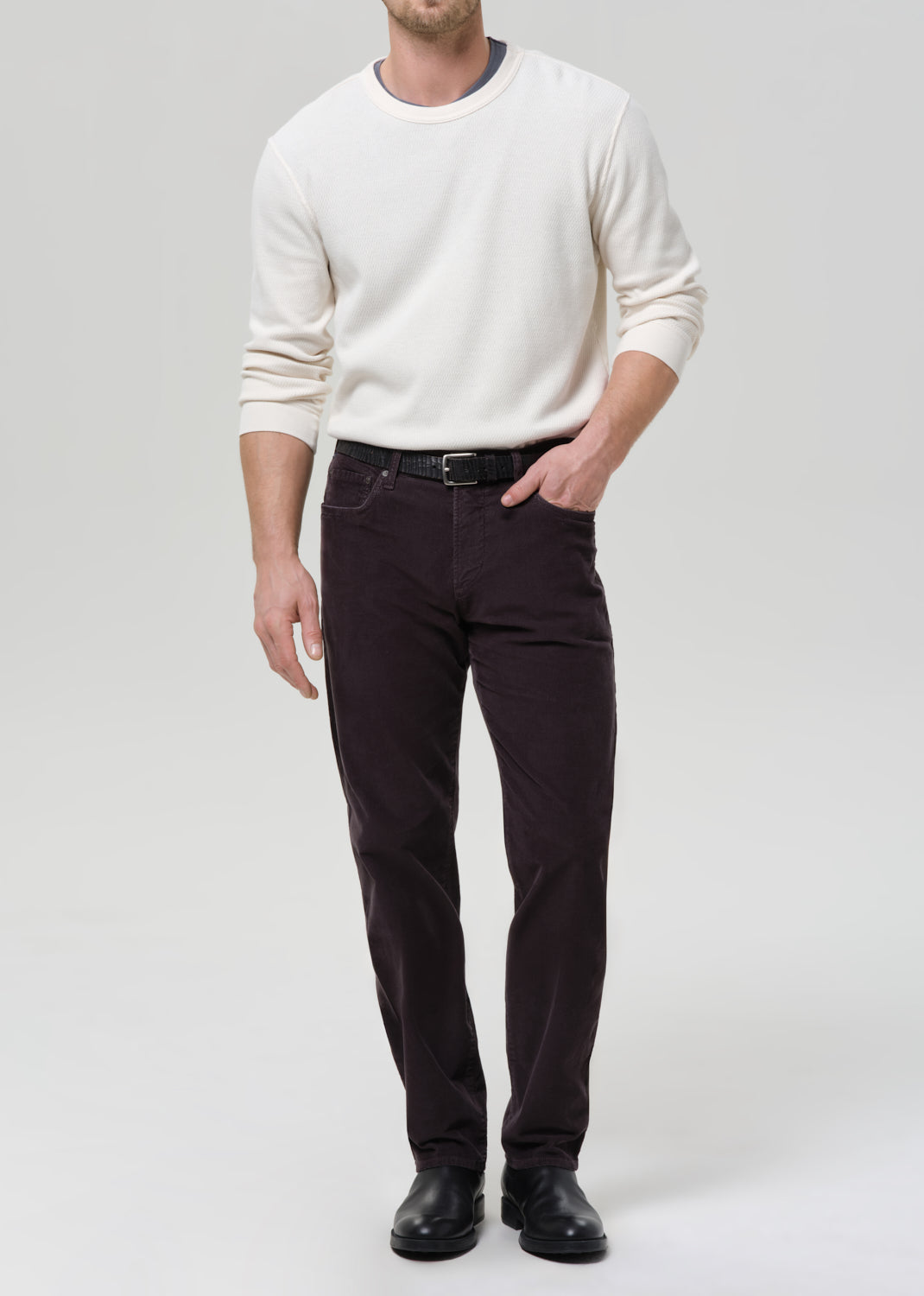 A man wearing a cream long-sleeve crewneck sweater, dark brown slim-fit trousers, a black belt, and black leather shoes, standing against a grey background.