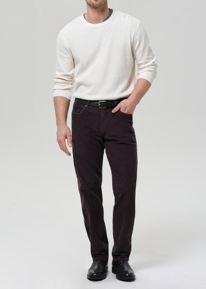 A man wearing a cream long-sleeve crewneck sweater, dark brown slim-fit trousers, a black belt, and black leather shoes, standing against a grey background.