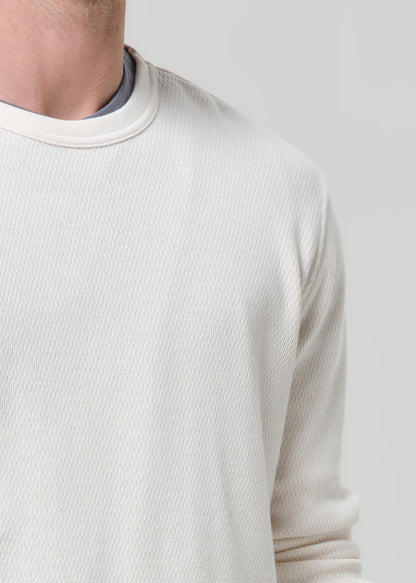 Close-up of a person wearing a textured off-white crewneck sweater, showing the ribbed collar and fabric texture against a grey background.