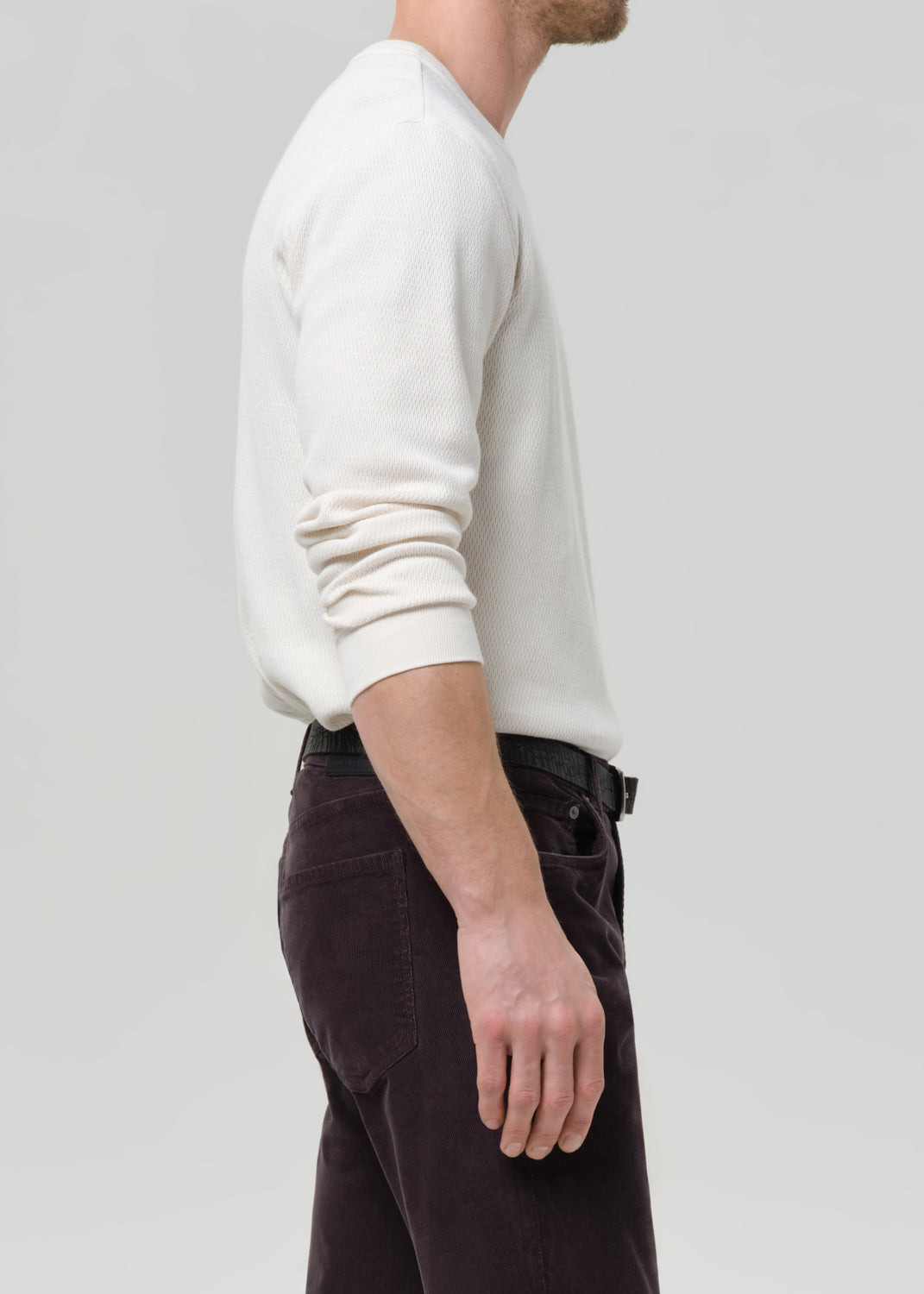 A man shown from the side wearing a cream-colored long-sleeve knit sweater and dark brown pants, photographed against a grey background.