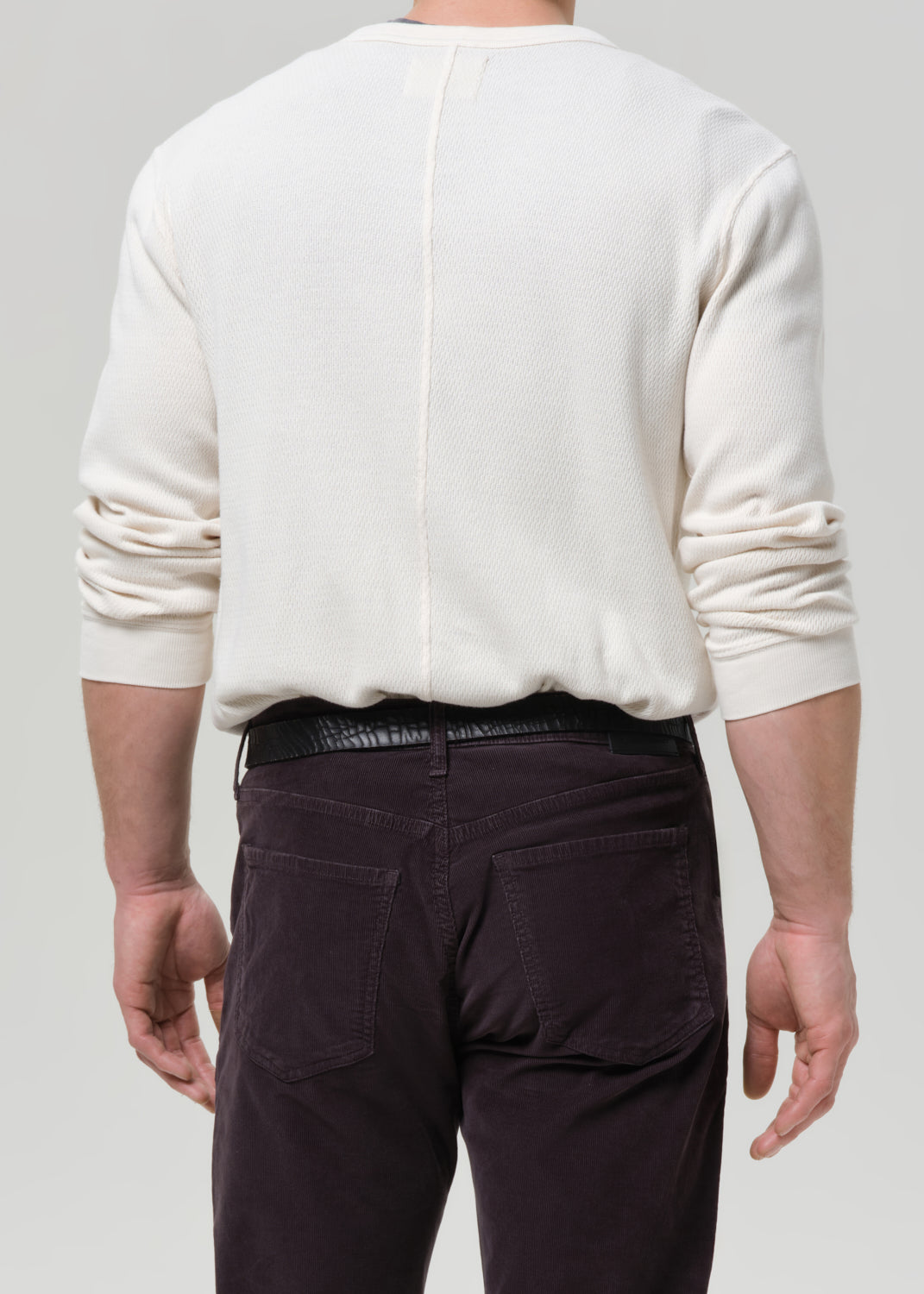 Back view of a man wearing an off-white long-sleeve knit sweater and dark brown pants, shown against a grey background.