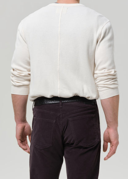 Back view of a man wearing an off-white long-sleeve knit sweater and dark brown pants, shown against a grey background.