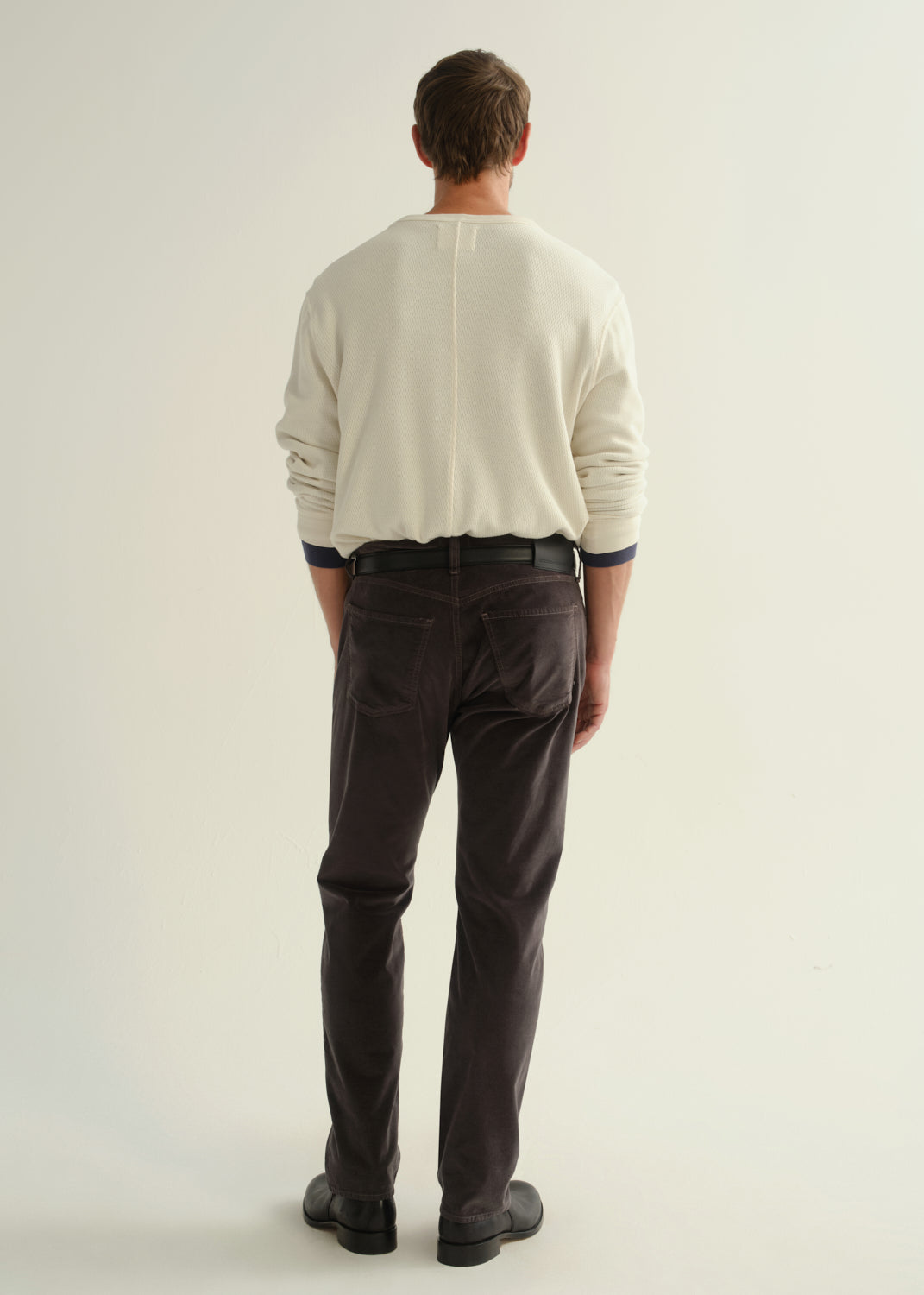 Person wearing a cream long sleeve crewneck sweater, dark brown pants, and black boots, back full body view on a light grey background.
