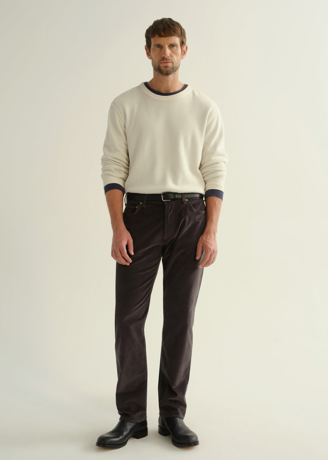 Person wearing a cream long sleeve crewneck sweater, dark brown pants, and black boots, front full body view on a light grey background.