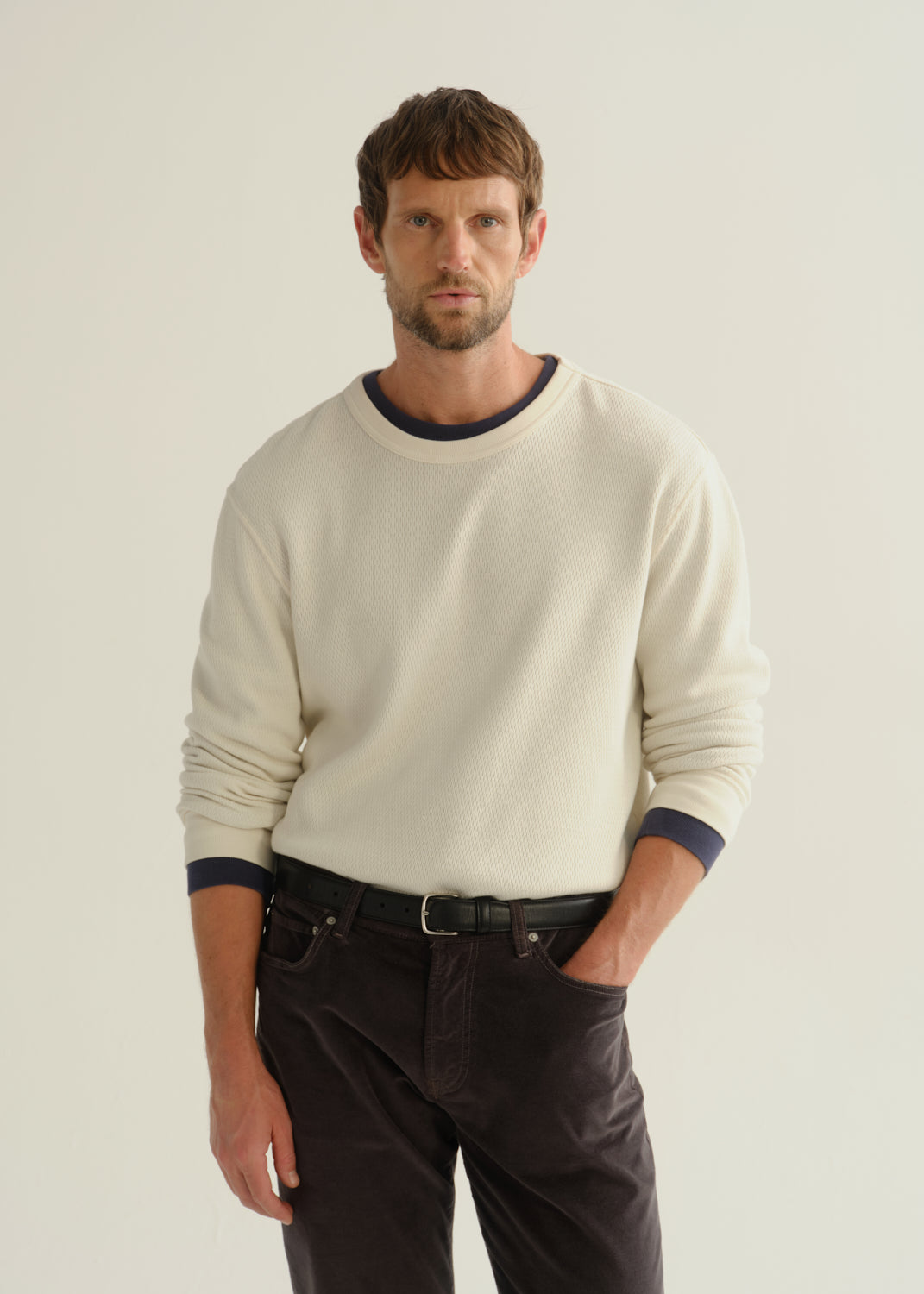 Person wearing a cream long sleeve crewneck sweater and dark brown pants, front cropped view on a light grey background.