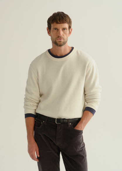 Person wearing a cream long sleeve crewneck sweater and dark brown pants, front cropped view on a light grey background.