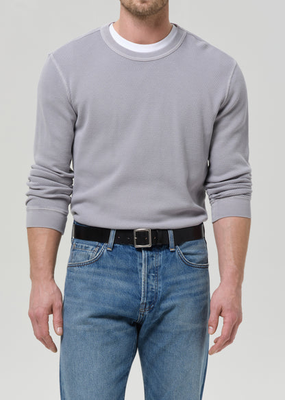A man wearing a light grey crewneck sweatshirt over a white undershirt, paired with blue jeans and a black belt, shown from the waist up against a grey background.