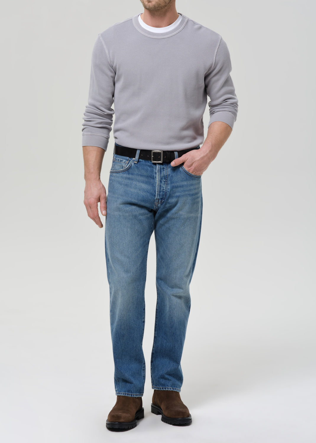 A man wearing a light grey long-sleeve crewneck sweater, blue jeans, a black belt, and brown boots, standing against a grey background.