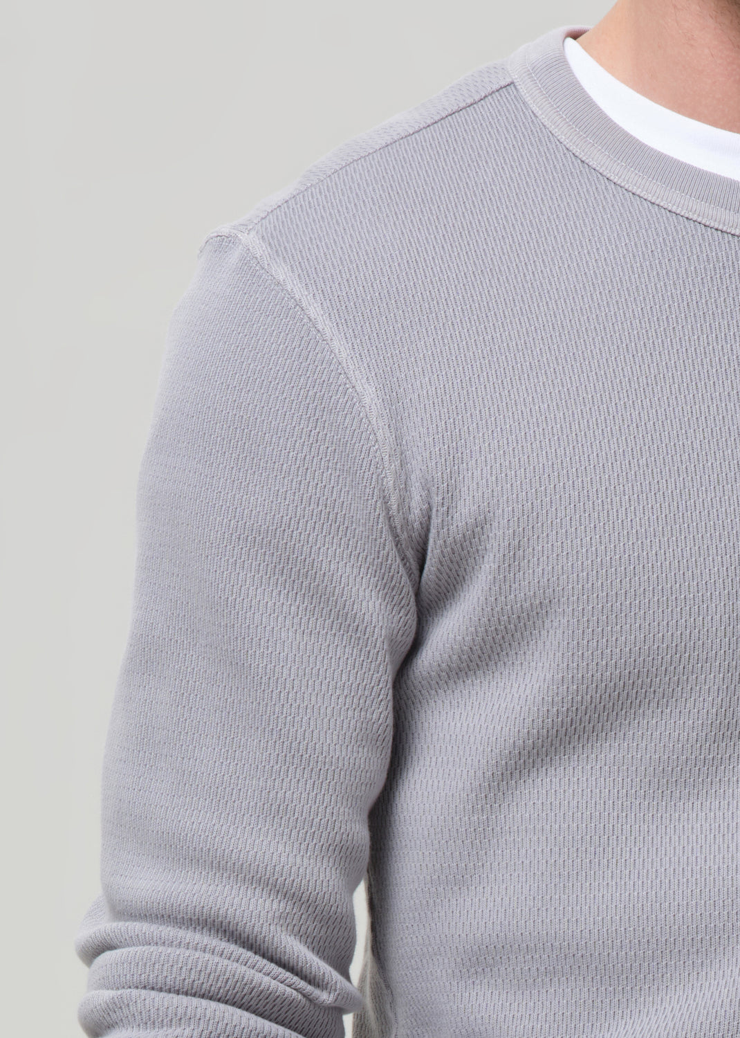 Close-up of a person wearing a light grey ribbed knit crewneck sweater over a white undershirt, showing the textured fabric and shoulder seam.