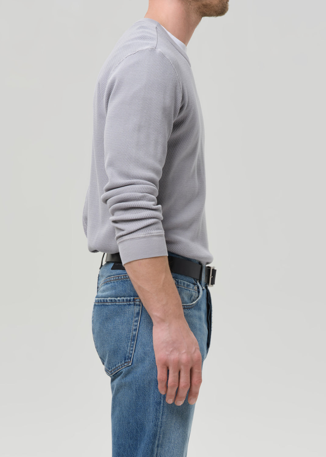 A man shown in side profile wearing a light grey long-sleeve crewneck sweater, blue jeans, and a black belt, against a grey background.