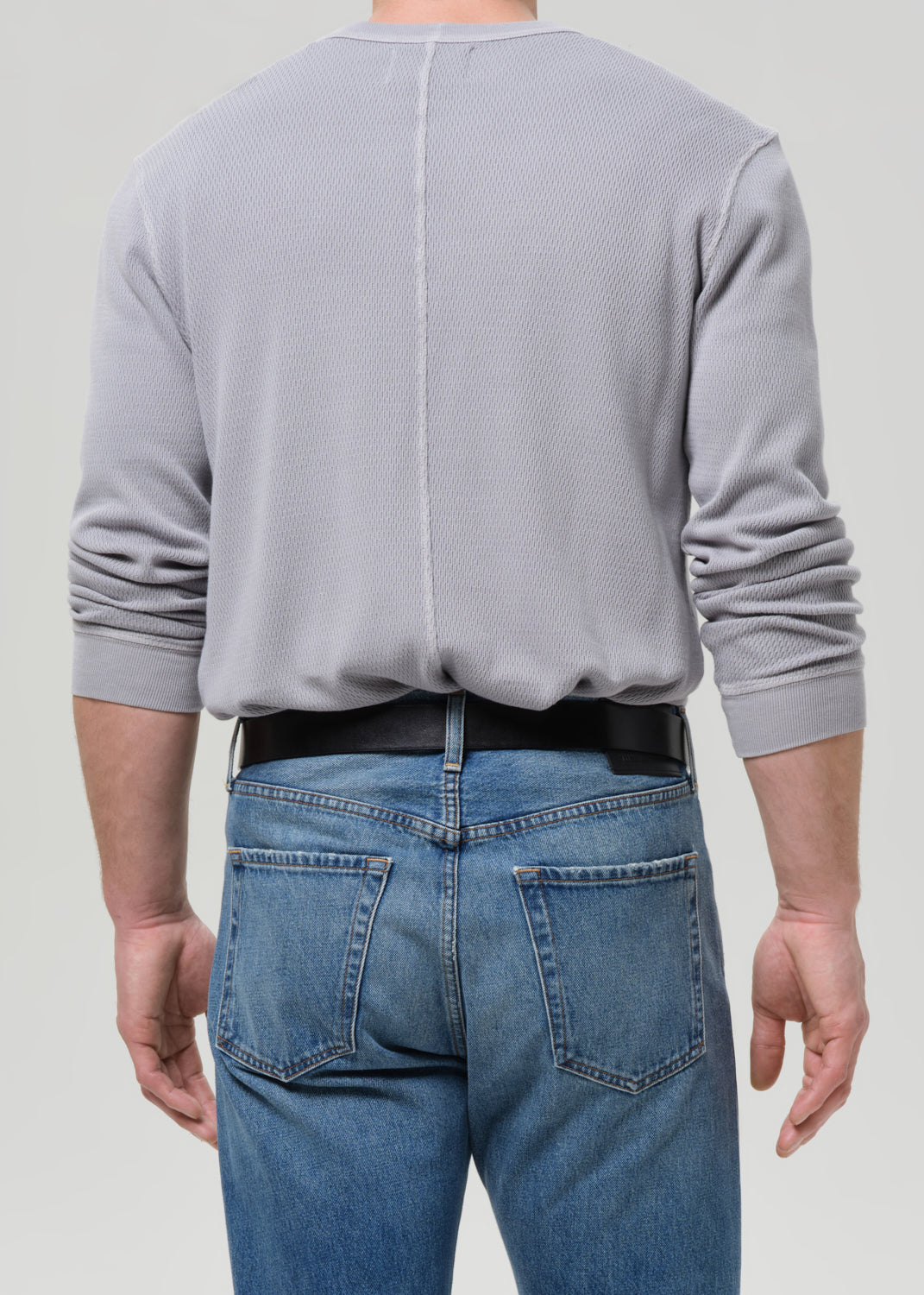 A man viewed from the back wearing a light grey ribbed long-sleeve shirt tucked into blue jeans with a black belt, against a grey background.