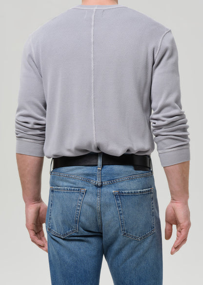 A man viewed from the back wearing a light grey ribbed long-sleeve shirt tucked into blue jeans with a black belt, against a grey background.
