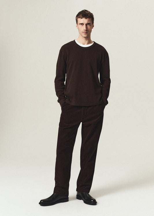 Man wearing a dark brown waffle crewneck top layered over a white crewneck tee with dark brown pants on a grey background.