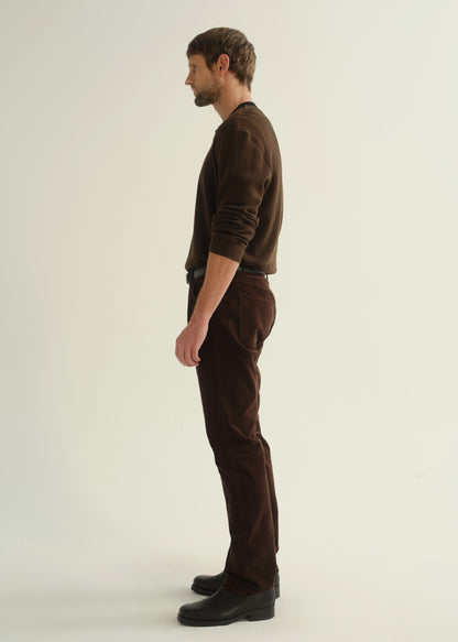 Person wearing a dark brown crewneck sweater, matching dark brown pants, and black boots, side full body view on a light grey background.