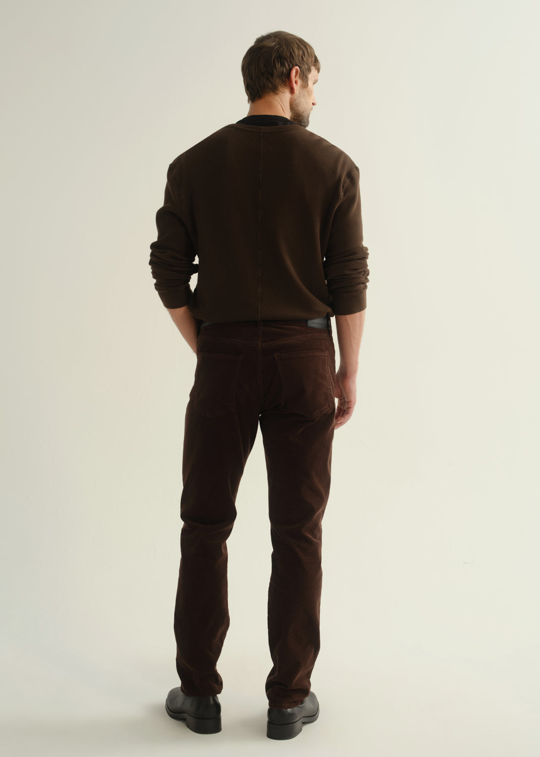 Person wearing a dark brown crewneck sweater, matching dark brown pants, and black boots, back full body view on a light grey background.