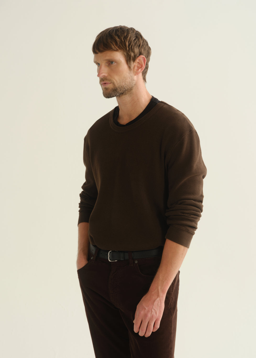 Person wearing a dark brown crewneck sweater and dark brown pants, angled front cropped view on a light grey background.
