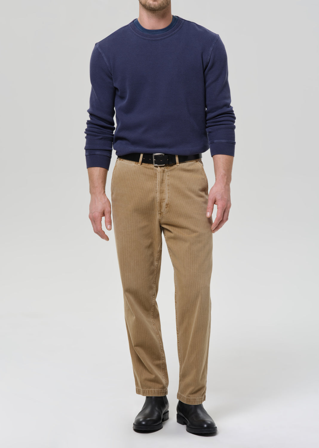 A man wearing a navy blue long-sleeve crewneck sweater, tan corduroy trousers, a black belt, and black leather shoes, standing against a grey background.