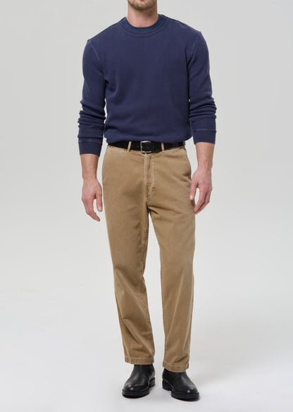 A man wearing a navy blue long-sleeve crewneck sweater, tan corduroy trousers, a black belt, and black leather shoes, standing against a grey background.
