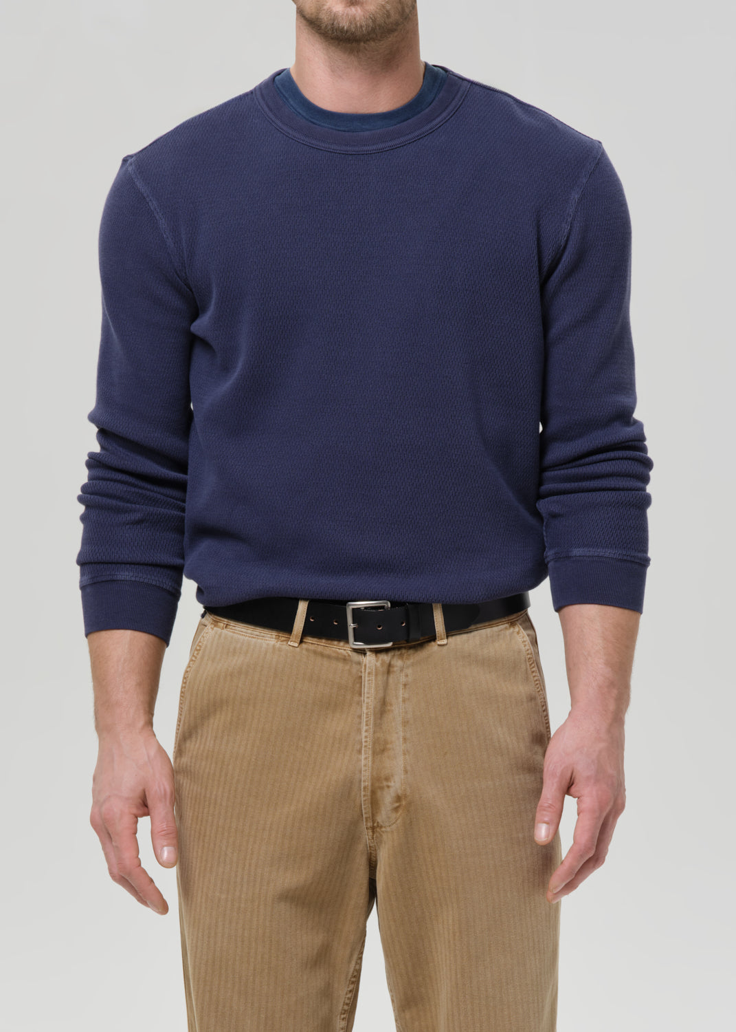 A man wearing a navy blue long-sleeve crewneck sweatshirt, paired with tan corduroy pants and a black belt, against a grey background.