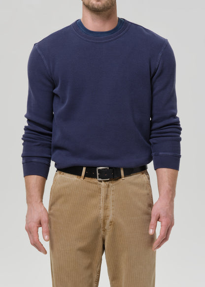 A man wearing a navy blue long-sleeve crewneck sweatshirt, paired with tan corduroy pants and a black belt, against a grey background.