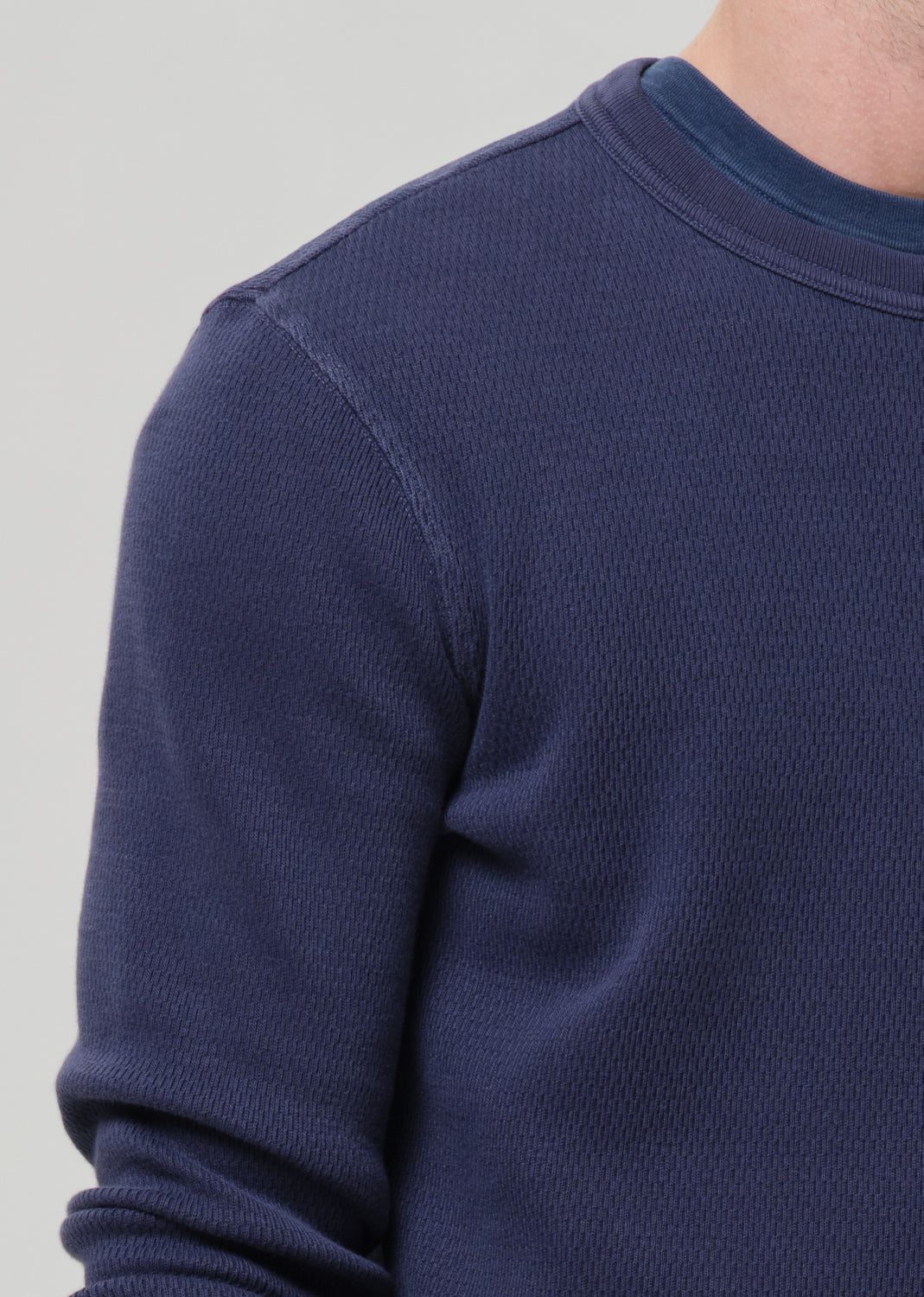 Close-up of a person wearing a textured navy blue long-sleeve crewneck sweater, showing the ribbed collar and shoulder detail against a grey background.