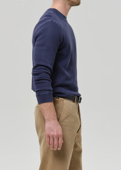 A man shown in side profile wearing a navy blue long-sleeve crewneck sweater and tan corduroy trousers against a grey background.
