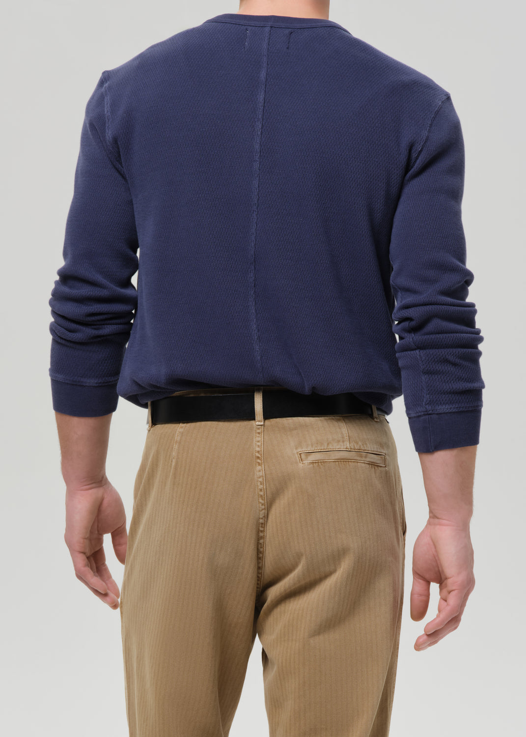 A man viewed from the back wearing a navy blue long-sleeve crewneck sweatshirt and tan corduroy trousers, paired with a black belt, against a grey background.