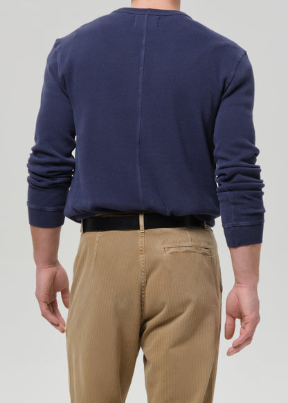 A man viewed from the back wearing a navy blue long-sleeve crewneck sweatshirt and tan corduroy trousers, paired with a black belt, against a grey background.