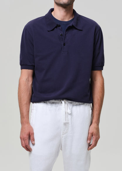 Man wearing a navy polo shirt and white pants against a plain background