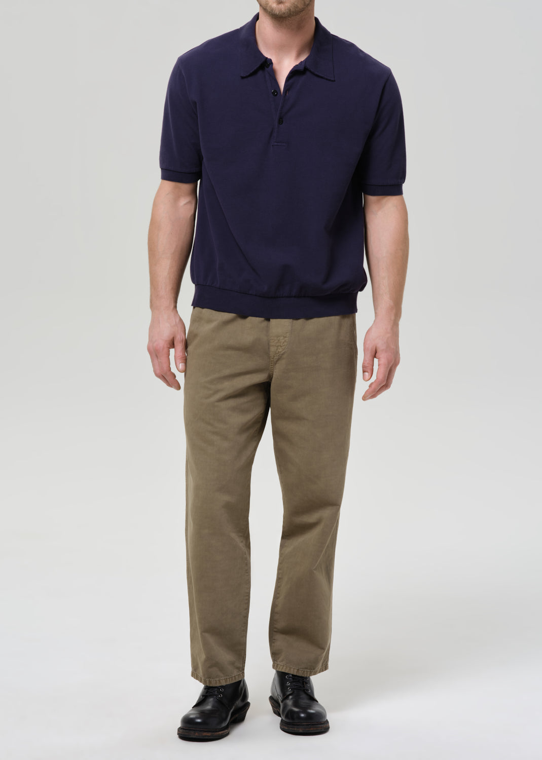 A man wearing a navy blue short-sleeve polo shirt, olive brown trousers, and black leather shoes, standing against a grey background.