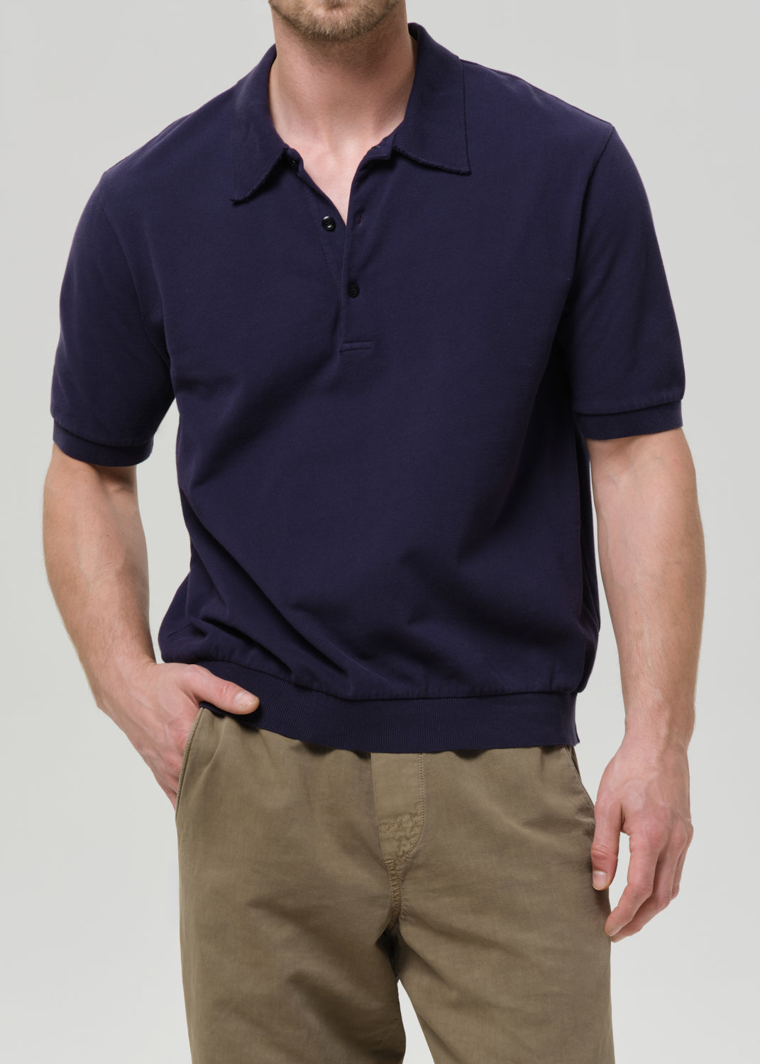 A man wearing a dark navy blue short-sleeve polo shirt and khaki pants, cropped from the chin down against a grey background.