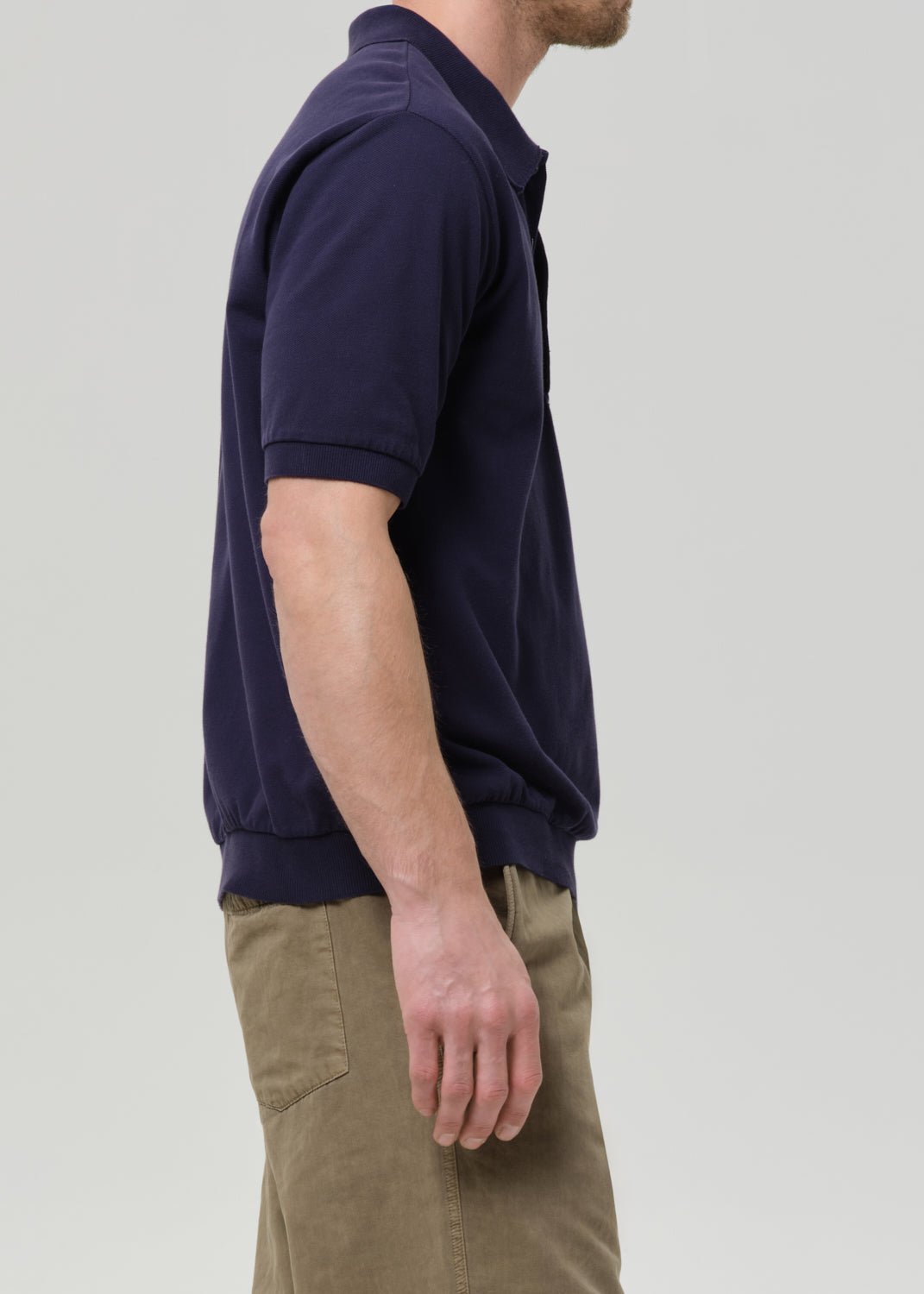 A man shown in side profile wearing a dark navy blue short-sleeve polo shirt and khaki pants, photographed against a grey background.
