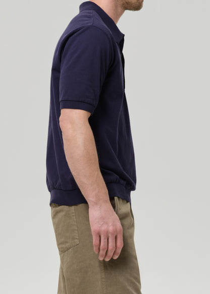 A man shown in side profile wearing a dark navy blue short-sleeve polo shirt and khaki pants, photographed against a grey background.
