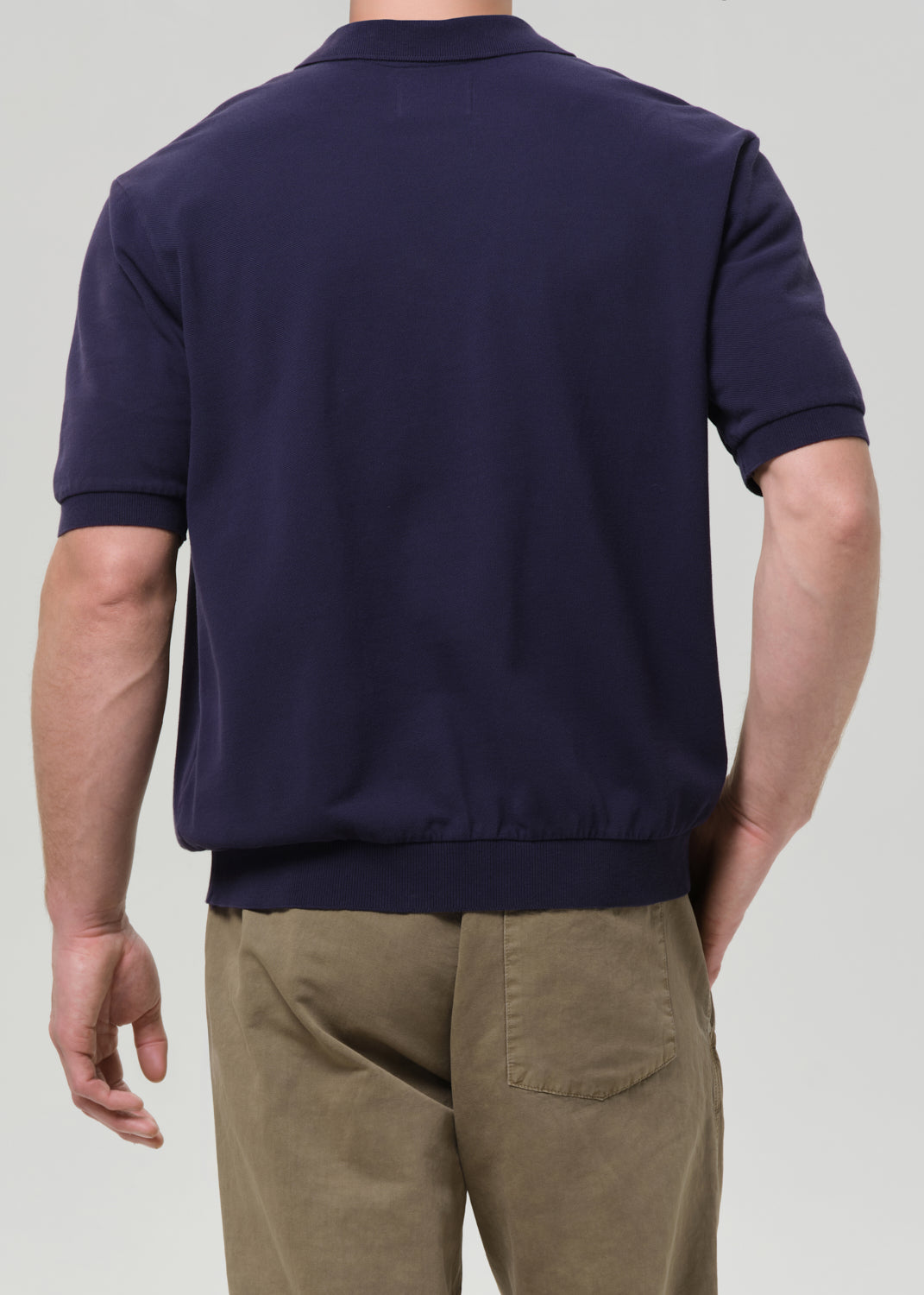 Rear view of a man wearing a navy blue short-sleeve knit polo shirt and khaki pants against a grey background.