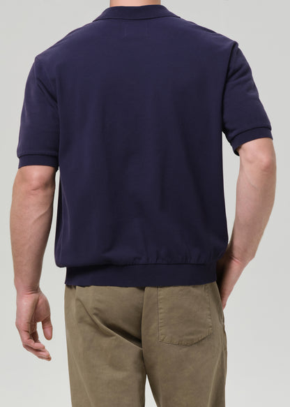 Rear view of a man wearing a navy blue short-sleeve knit polo shirt and khaki pants against a grey background.