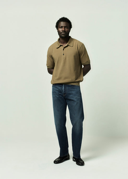 Man wearing a brown polo shirt and blue jeans on a plain background
