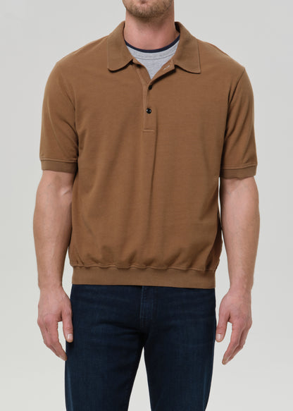 A man wearing a brown short-sleeve knit polo shirt over a gray t-shirt and dark blue jeans, shown from the chin down against a grey background.