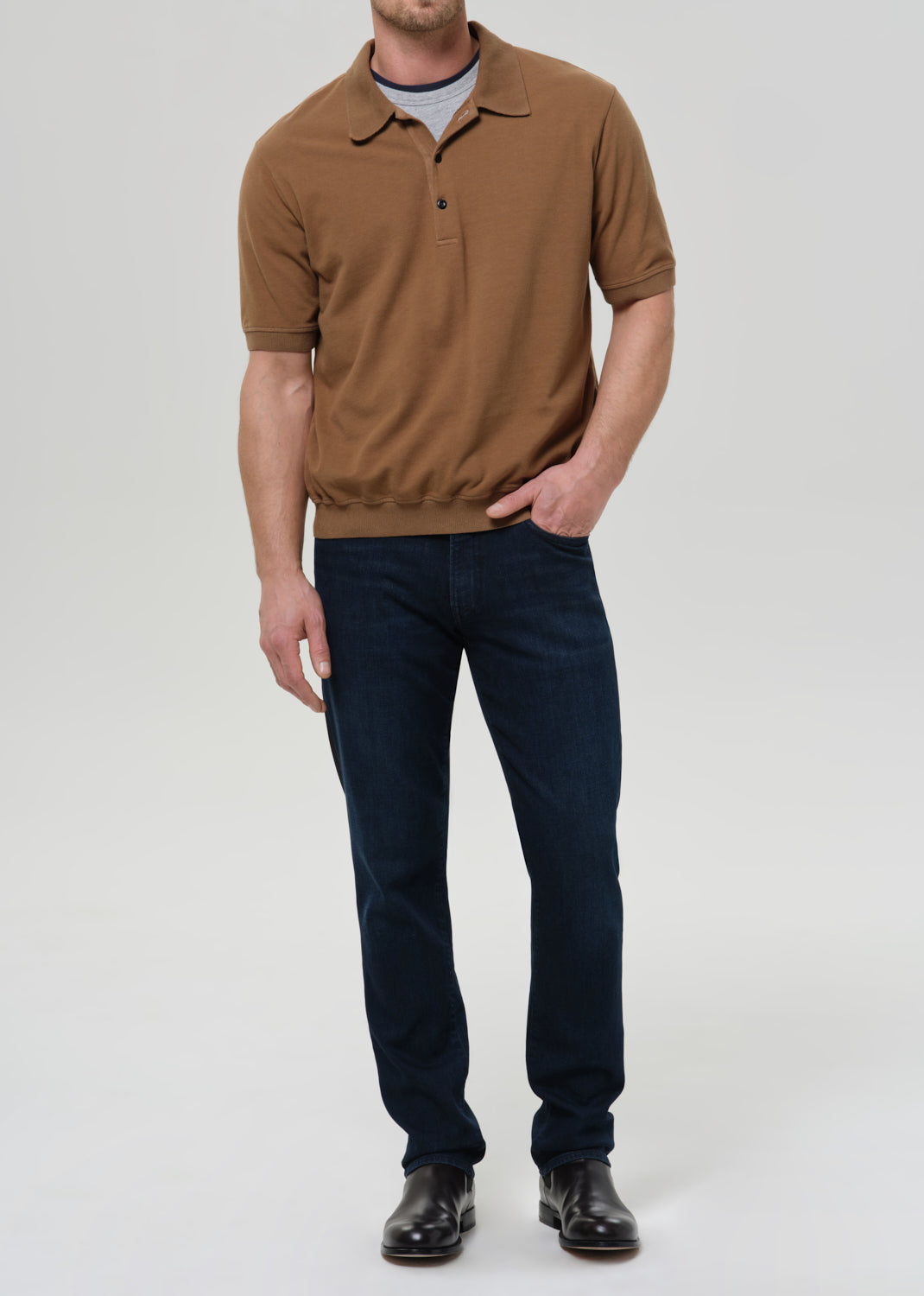 A man wearing a brown short-sleeve polo shirt, dark blue jeans, and black shoes, standing against a grey background.