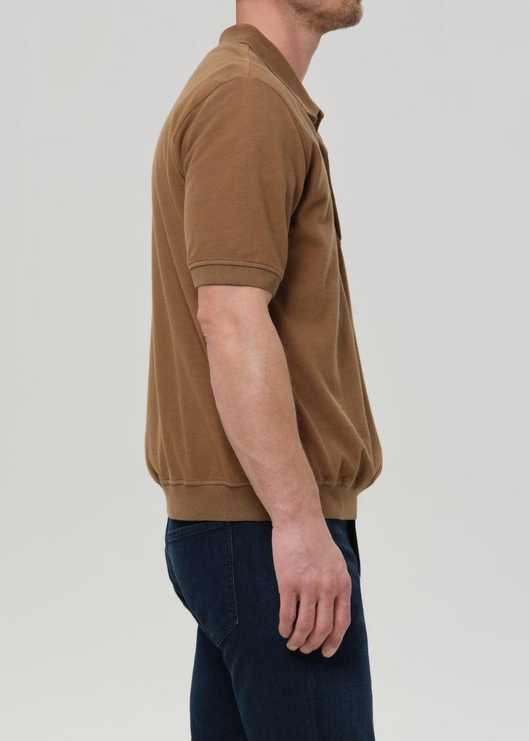 Side profile of a man wearing a brown short-sleeve polo shirt and dark blue jeans against a grey background.