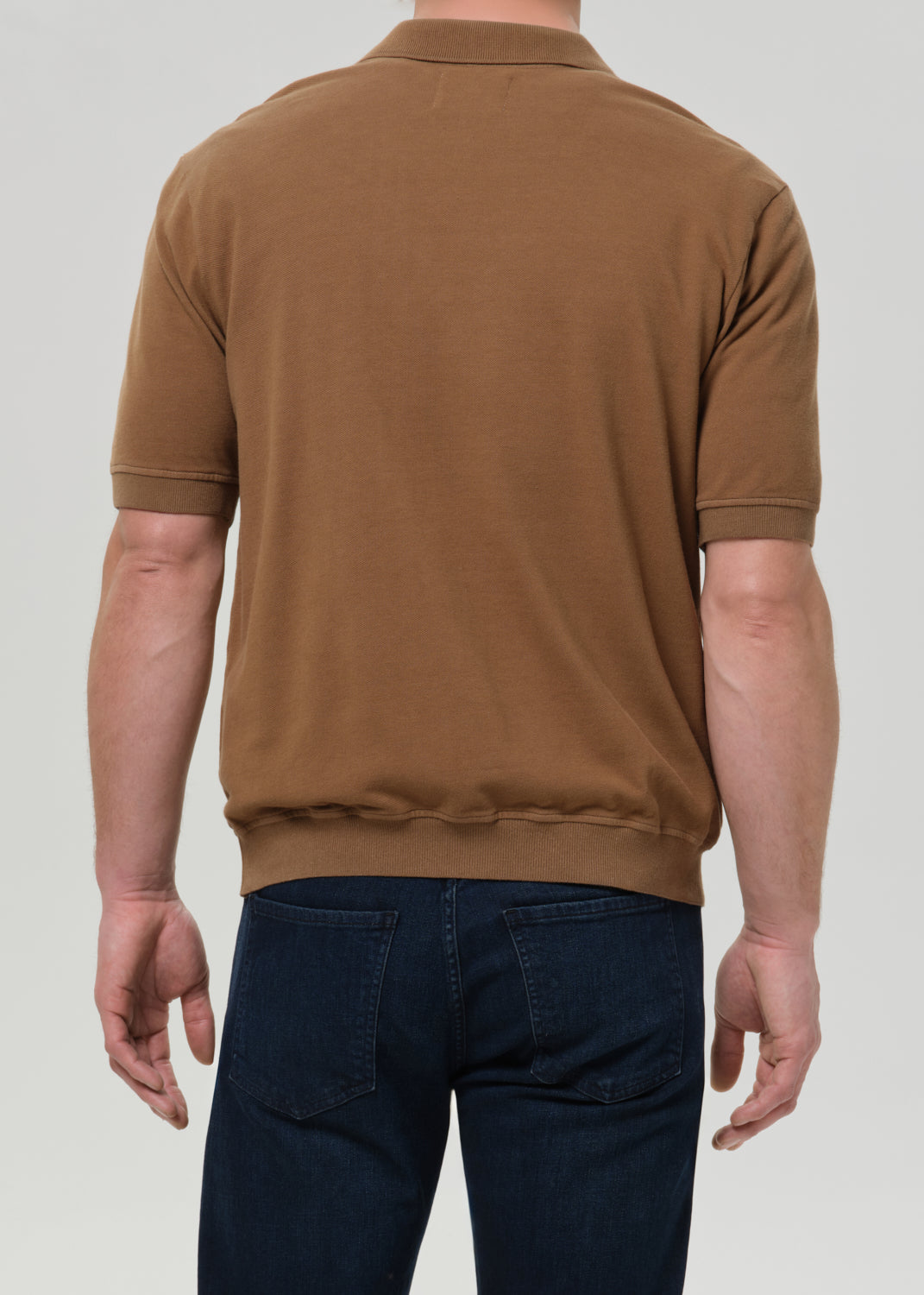 A man viewed from the back wearing a brown short-sleeve knit polo shirt and dark blue jeans, set against a grey background.