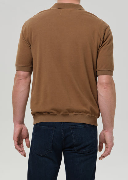 A man viewed from the back wearing a brown short-sleeve knit polo shirt and dark blue jeans, set against a grey background.